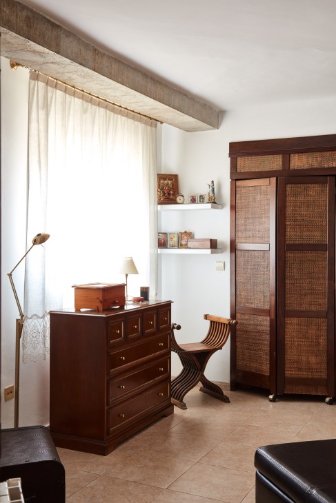Minimalist bedroom with wooden furniture, white shelving, and natural light from curtained window.