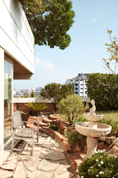 A sunny garden terrace with stone seating area, wooden chair, potted plants, and Espai Verd building visible in background.