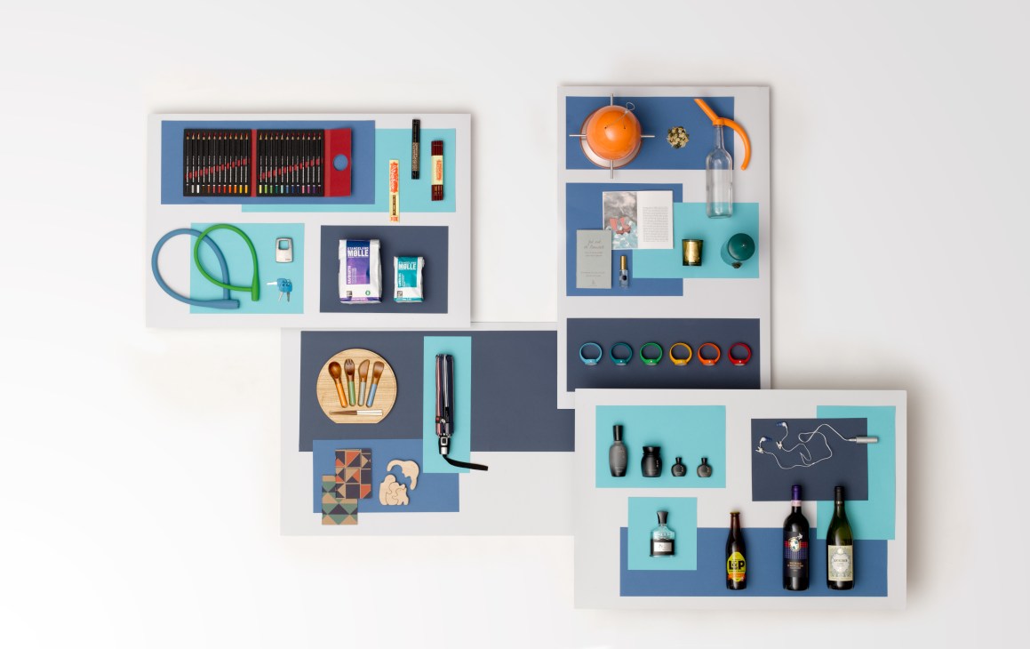 Flat lay arrangement of lifestyle products including art supplies, bike locks, notebooks, kitchenware, candles, wine bottles and accessories on blue and turquoise backgrounds.