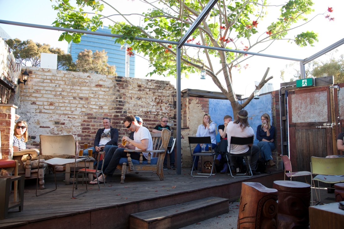 Outdoor courtyard seating area with brick walls, trees, and people gathering under a pergola at The Bird bar.
