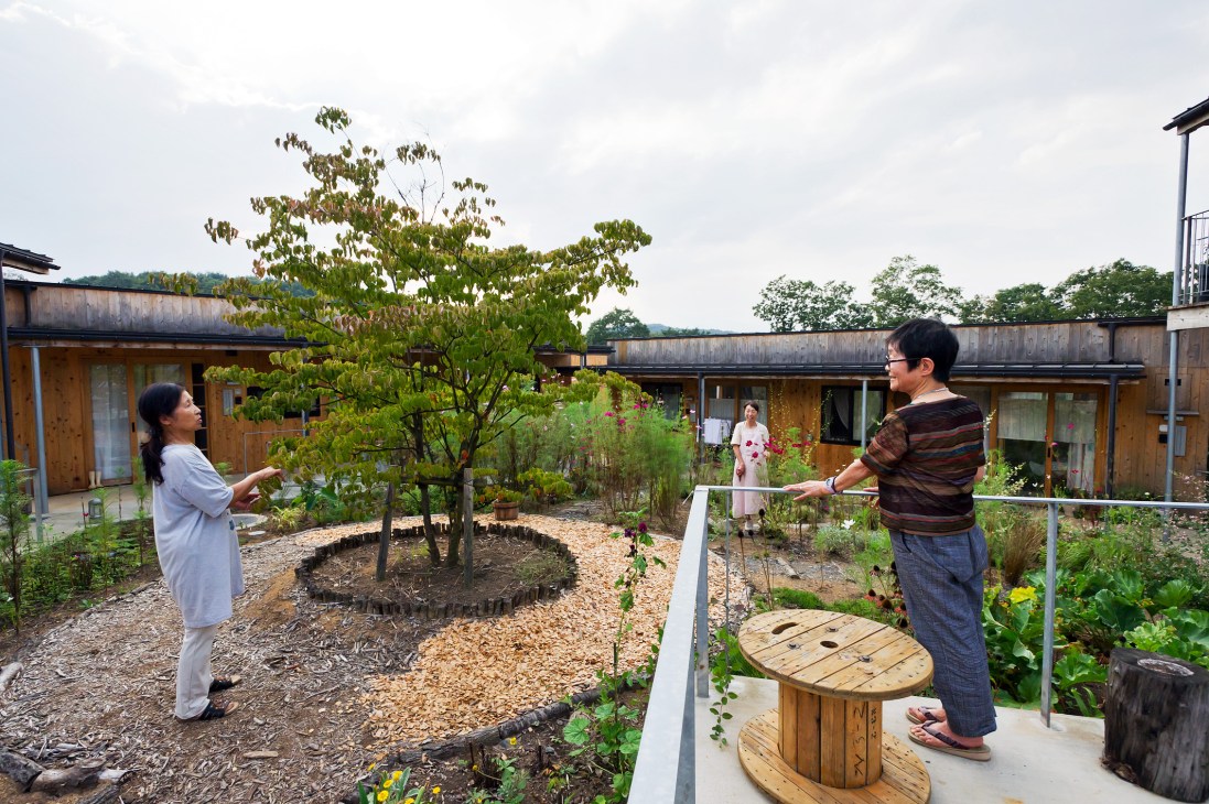 Two residents play cornhole in a shared courtyard surrounded by gardens and wooden buildings at Yuimarl Nasu.