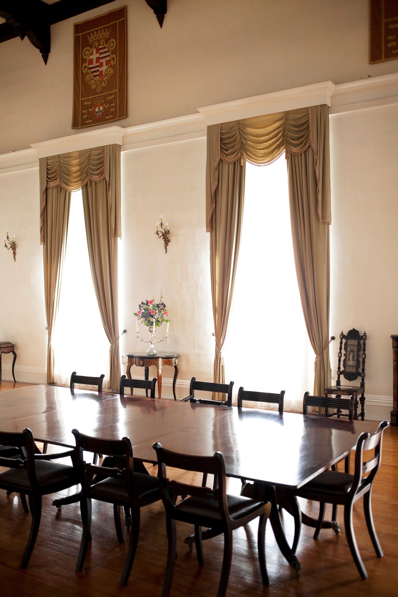 Formal dining room with long table, black chairs, tall windows with draped curtains, and ornate coat of arms on wall.