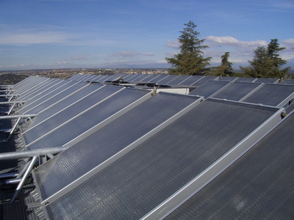 Rows of solar thermal collectors installed on a rooftop with trees and cityscape visible in the background.