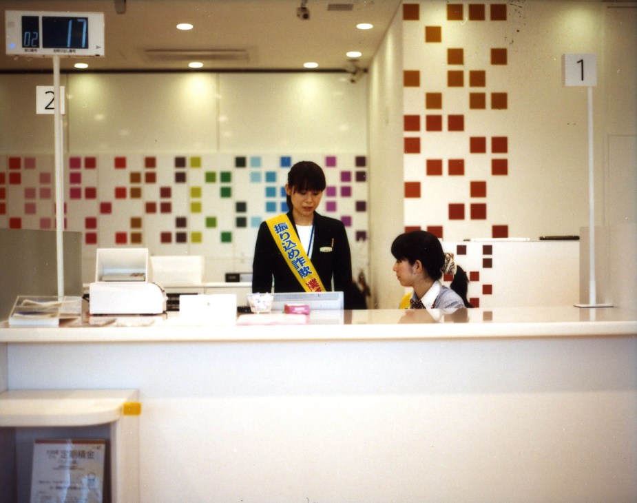 Bank teller at Sugamo Shinkin Bank's colorful Niiza branch with multicolored square tiles on the wall behind the counter.