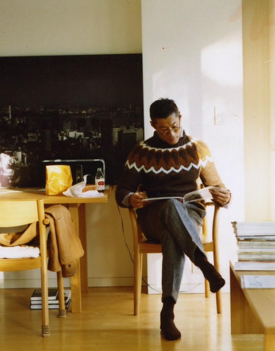 Man reading a magazine while seated in a wooden chair wearing a patterned knit jumper and glasses.