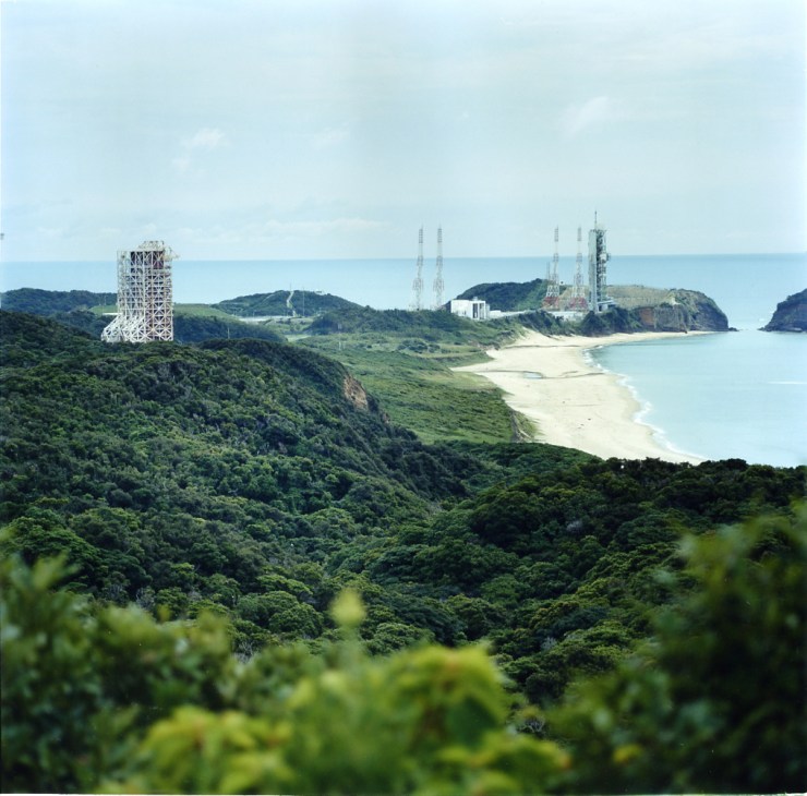 Yoshinobu launch complex with white service tower and communication antennas on a peninsula surrounded by dense green forest and turquoise ocean.