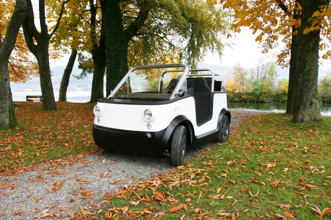 White and black E'mo electric car parked on an autumn leaf-covered path beside tall trees and a lake.