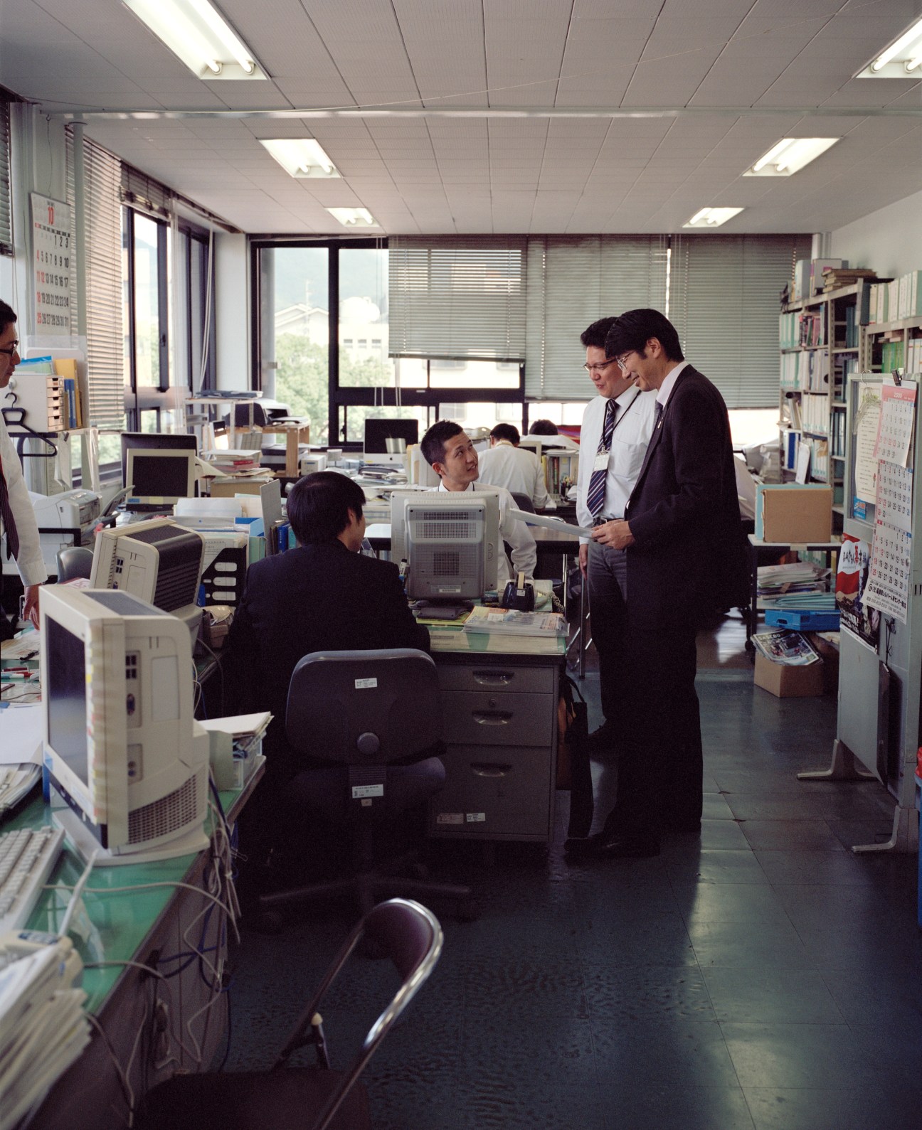 Tomihisa Taue chats with city hall staff in a busy office filled with computers and filing cabinets.