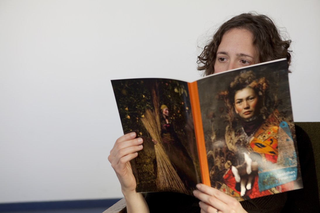 Woman with dark curly hair reading an open art book with orange spine and portrait imagery.