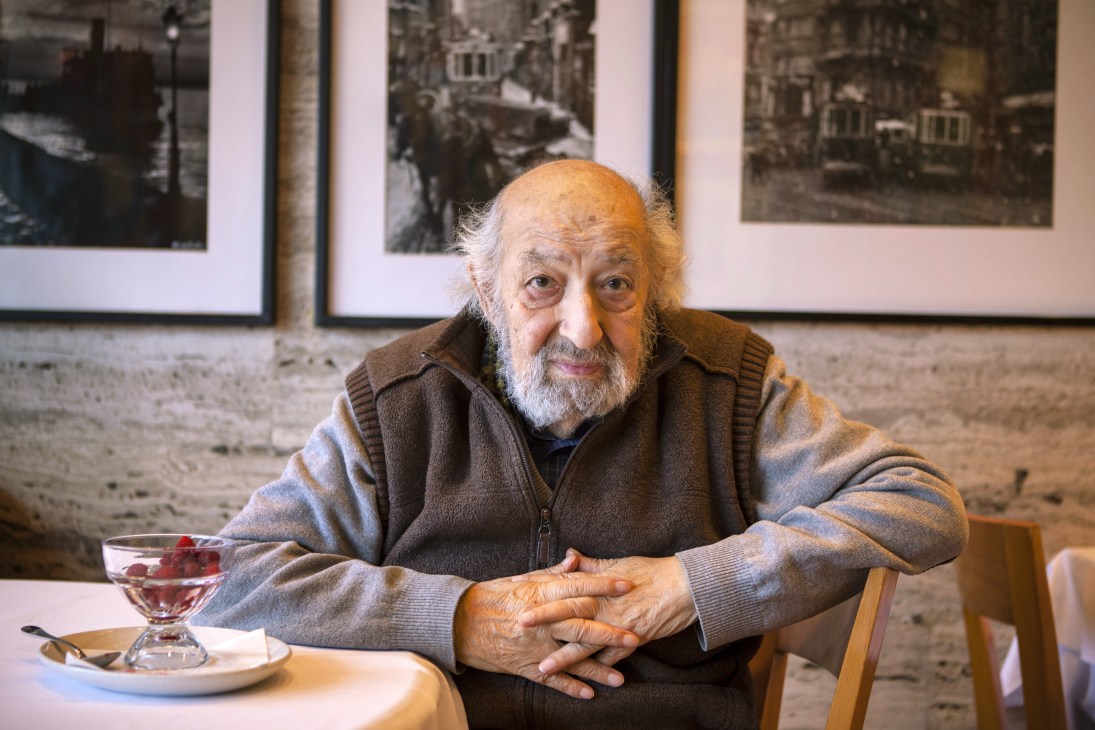 Ara Güler, an elderly man with white beard, seated at a restaurant table with framed photographs on the wall behind him.