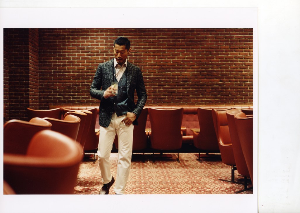 Man wearing Gucci jacket and Oliver Peoples glasses in retro 1970s bar with brick walls and red leather seating.