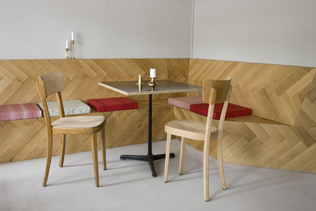 Café interior with wooden herringbone wall paneling, small table, and two light wood chairs with red and pink cushioned bench seating.