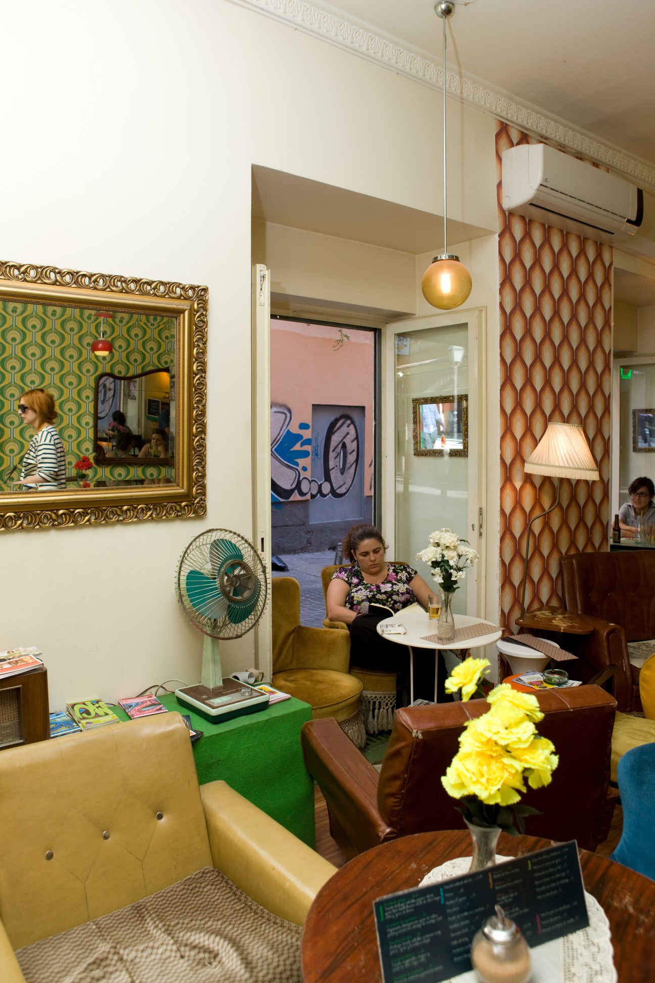 Interior of vintage-furnished café Lolina with 1950s-70s furniture, retro wallpaper, and a woman seated at a table.