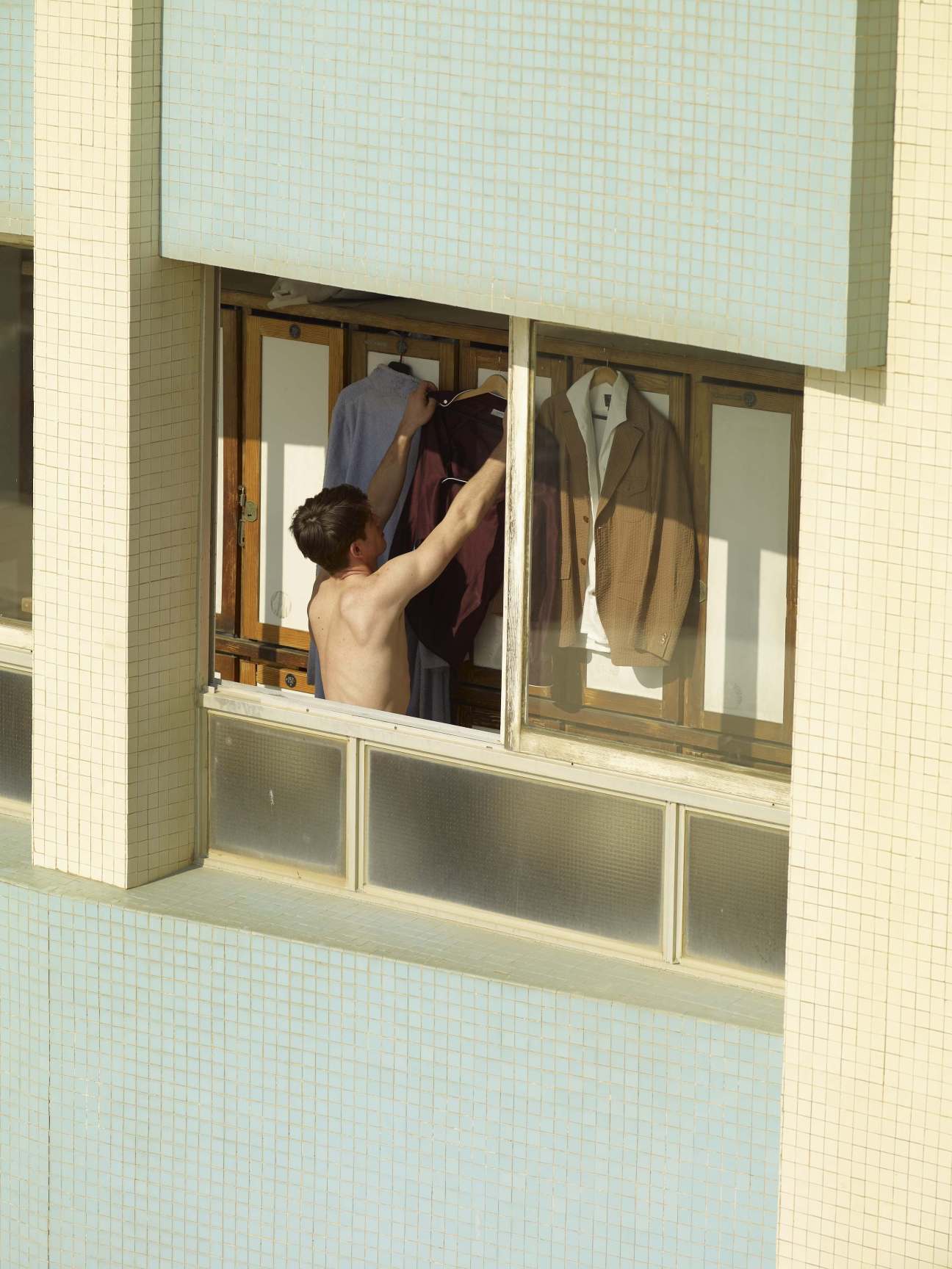 Shirtless man selecting clothing from hangers in a changing room window at a beach club.