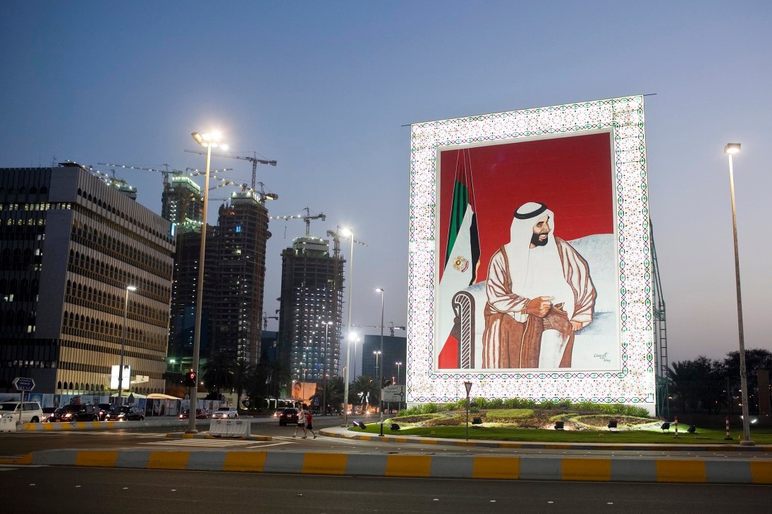 Large outdoor portrait of Sheikh Zayed on decorative background with Abu Dhabi skyline and construction cranes behind.