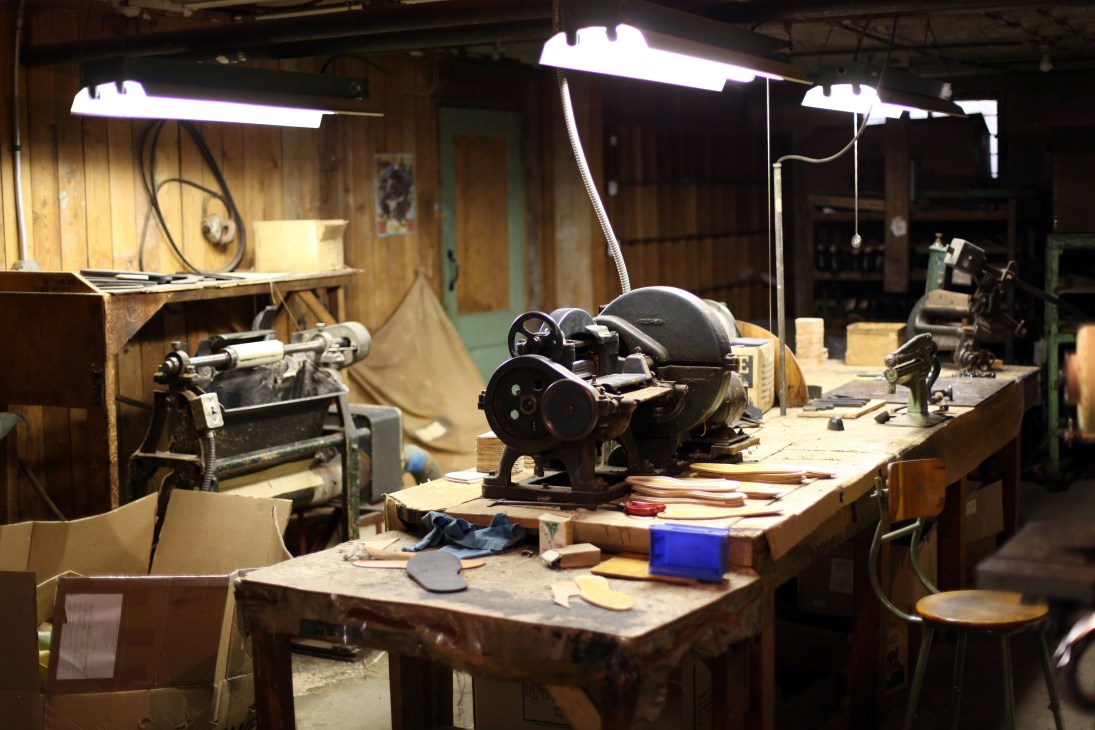Dayton Boots workshop with industrial workbenches, machinery, and leather materials under bright overhead lighting.