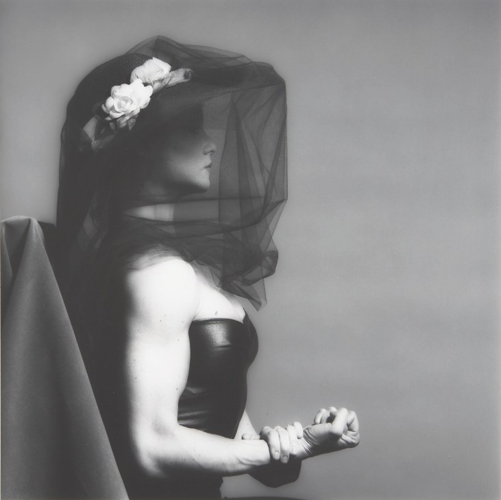 Black and white portrait photograph of a woman with muscular arms wearing a black strapless top and sheer veil with white flowers.