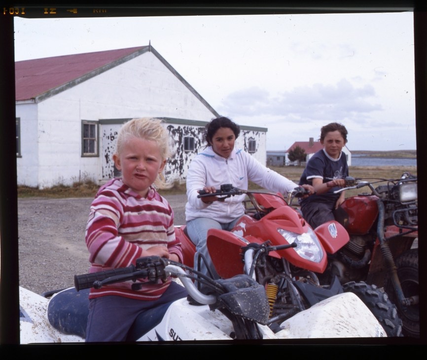 Three children sitting on red ATVs outside a house in Goose Green, Falkland Islands.