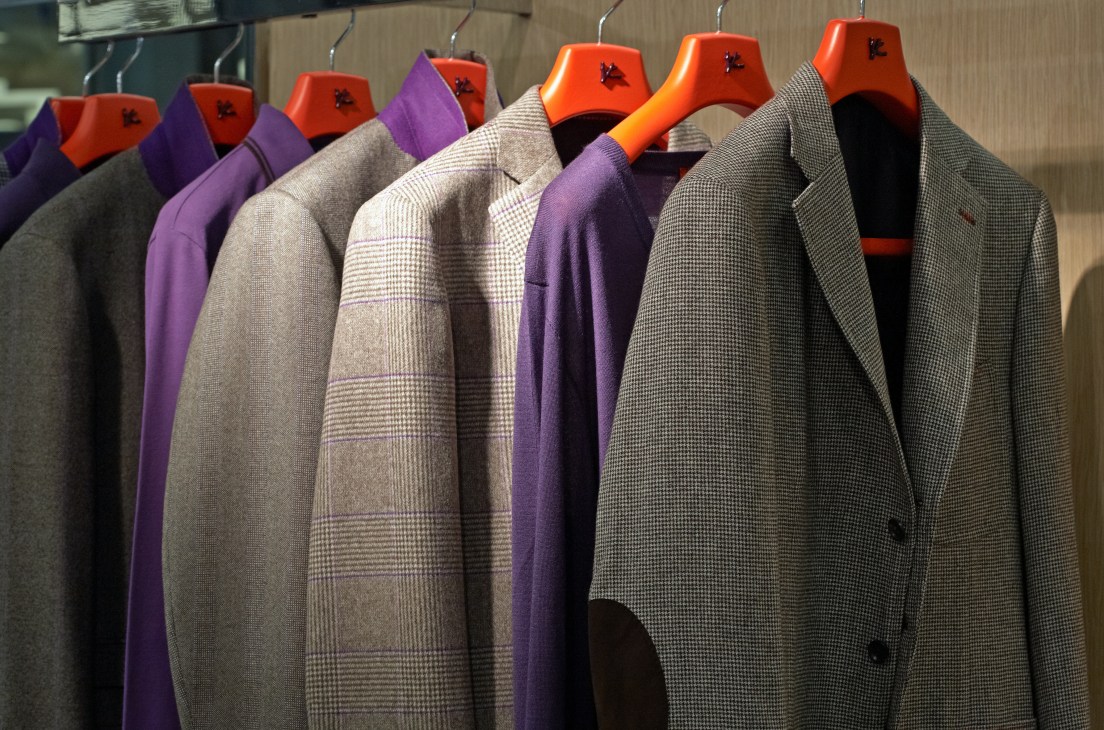 Menswear blazers and coats on hangers at Isaia's stand, featuring purple, checked, and neutral patterns with bright orange and red jacket forms.