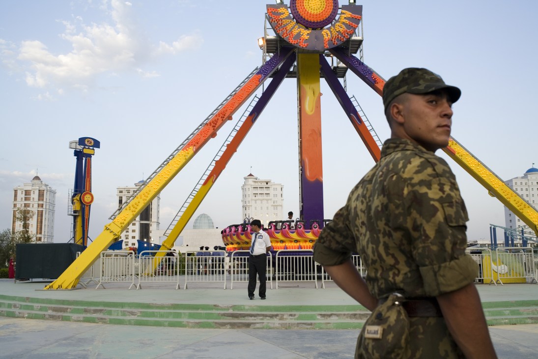 Army conscript in camouflage uniform stands before a large colorful fairground ride in Ashgabat.