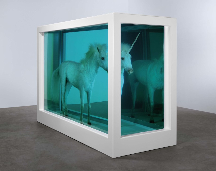 A white vitrine display case containing a taxidermied foal with a gold horn (unicorn) suspended in turquoise formaldehyde solution.