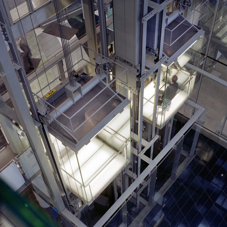 Overhead view of illuminated elevator shafts in the Post Tower, Bonn, showing multiple lift cars with metal frameworks and lighting.