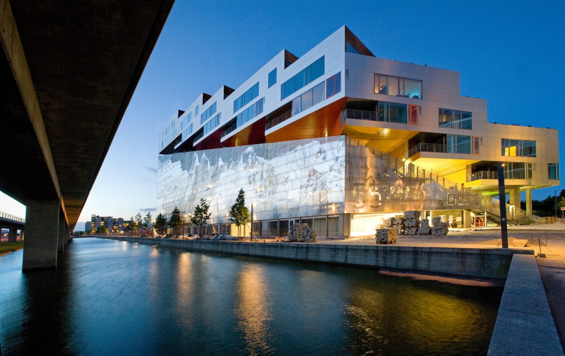 Mountain Dwellings, a cascading residential building with white facades and terracotta roof accents reflecting in water at dusk in Copenhagen.