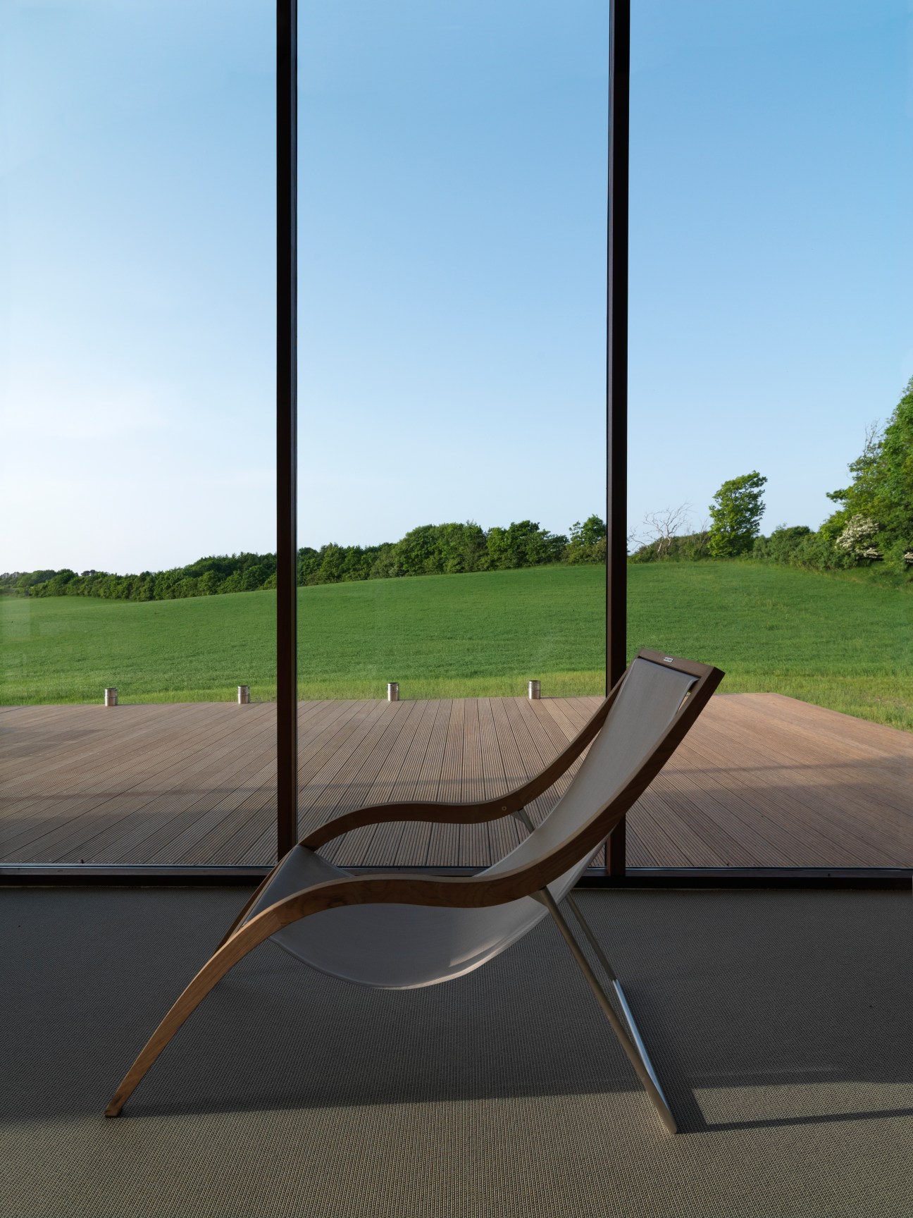 Modern curved lounge chair by floor-to-ceiling windows overlooking green fields and tree line.