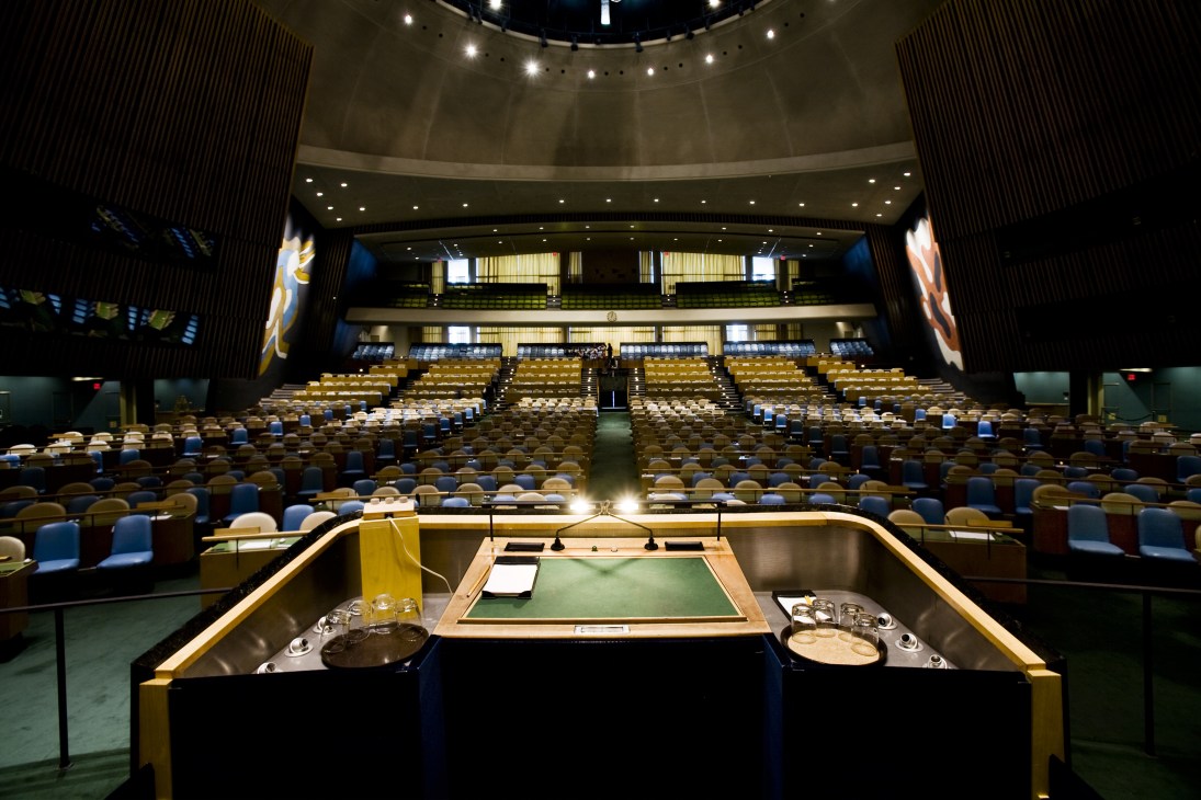 Empty UN General Assembly chamber with rows of beige and blue seats facing a raised platform with green surface under bright lighting.