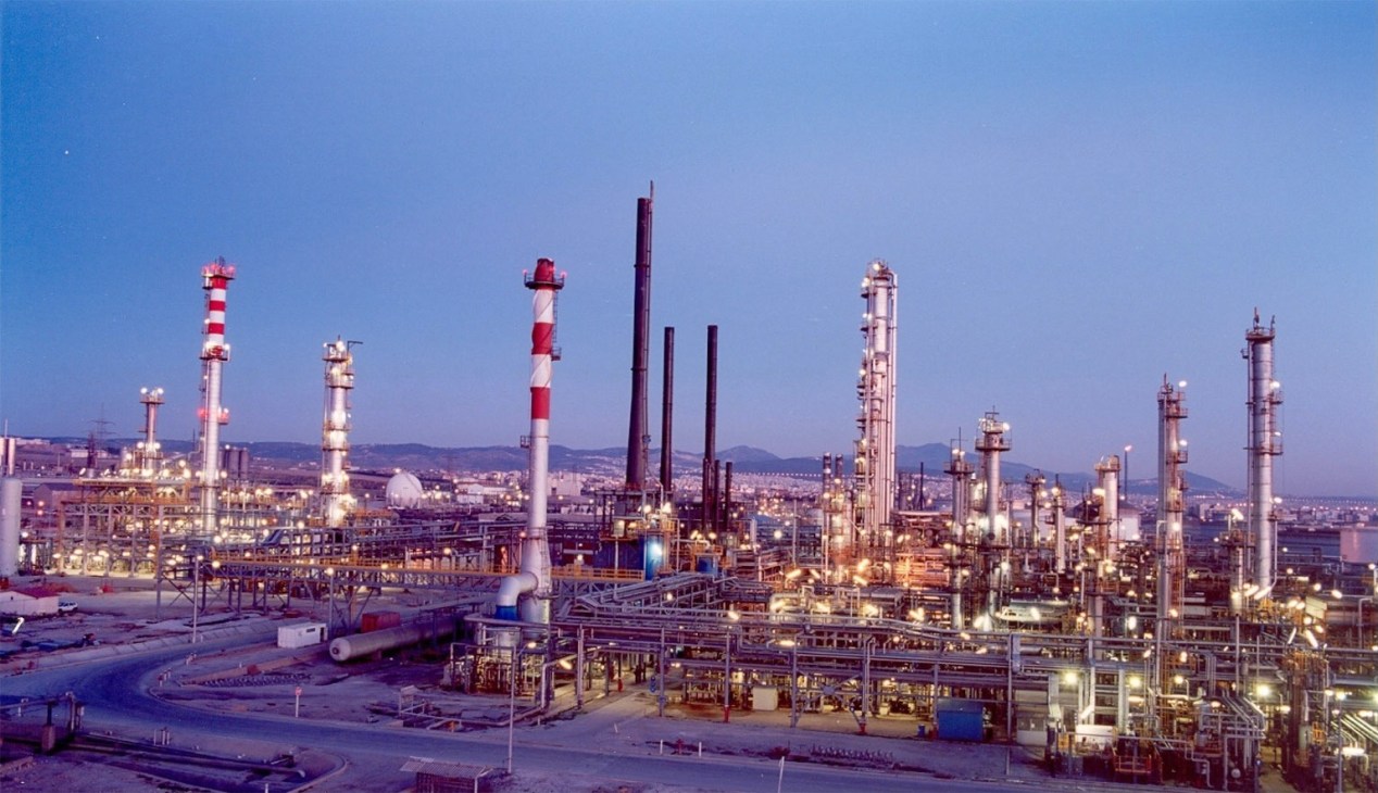 Hellenic Petroleum's oil refinery at dusk with illuminated towers and striped smokestacks.