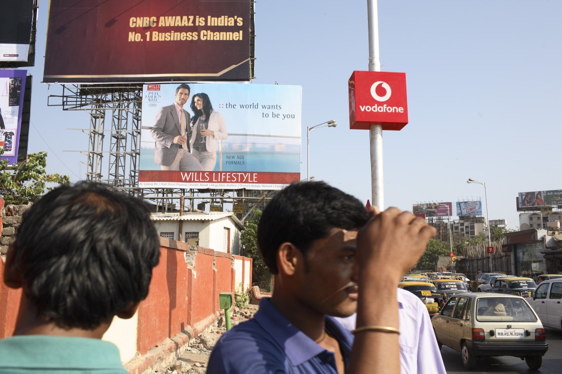 Mumbai street scene with multiple billboards including Vodafone and Wills Lifestyle ads above traffic and pedestrians.