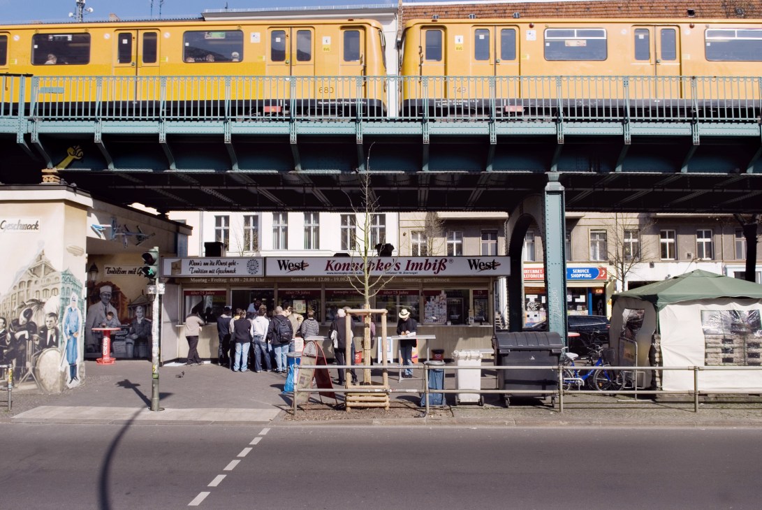 Konnopke's wurst stand beneath the yellow U2 U-Bahn railway arches with customers queuing outside.