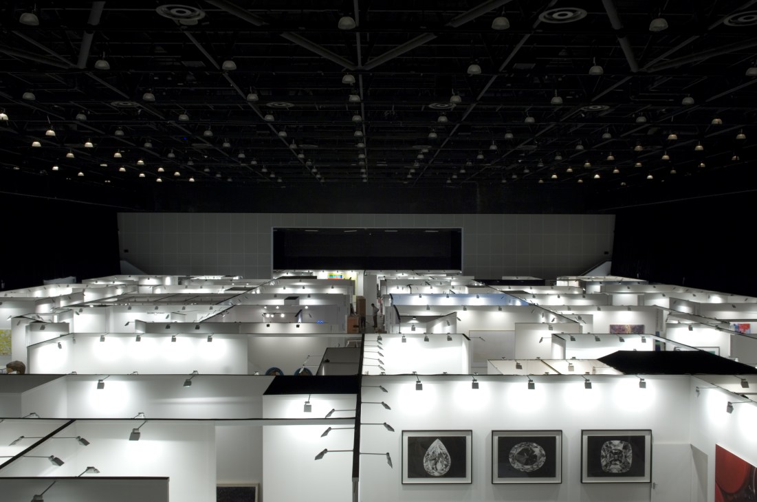 Overhead view of Art Dubai fair showing white gallery booths with track lighting in a large arena space.