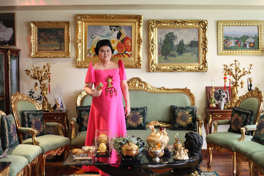 Imelda Marcos in a pink dress standing in her ornate living room with gold-framed artwork and decorative sculptures.