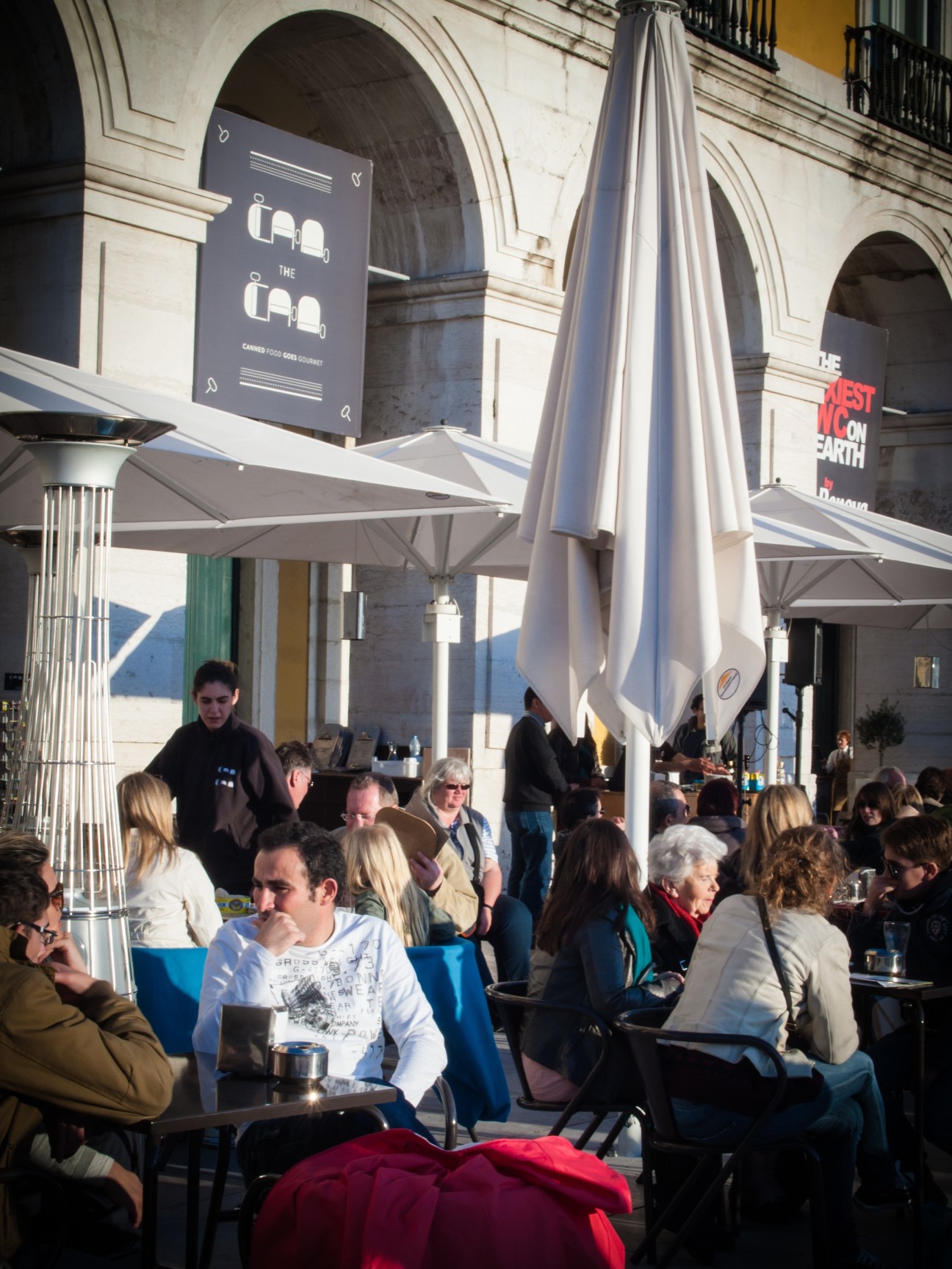 Outdoor café terrace with diners under white umbrellas in front of classical architecture with arched doorways.