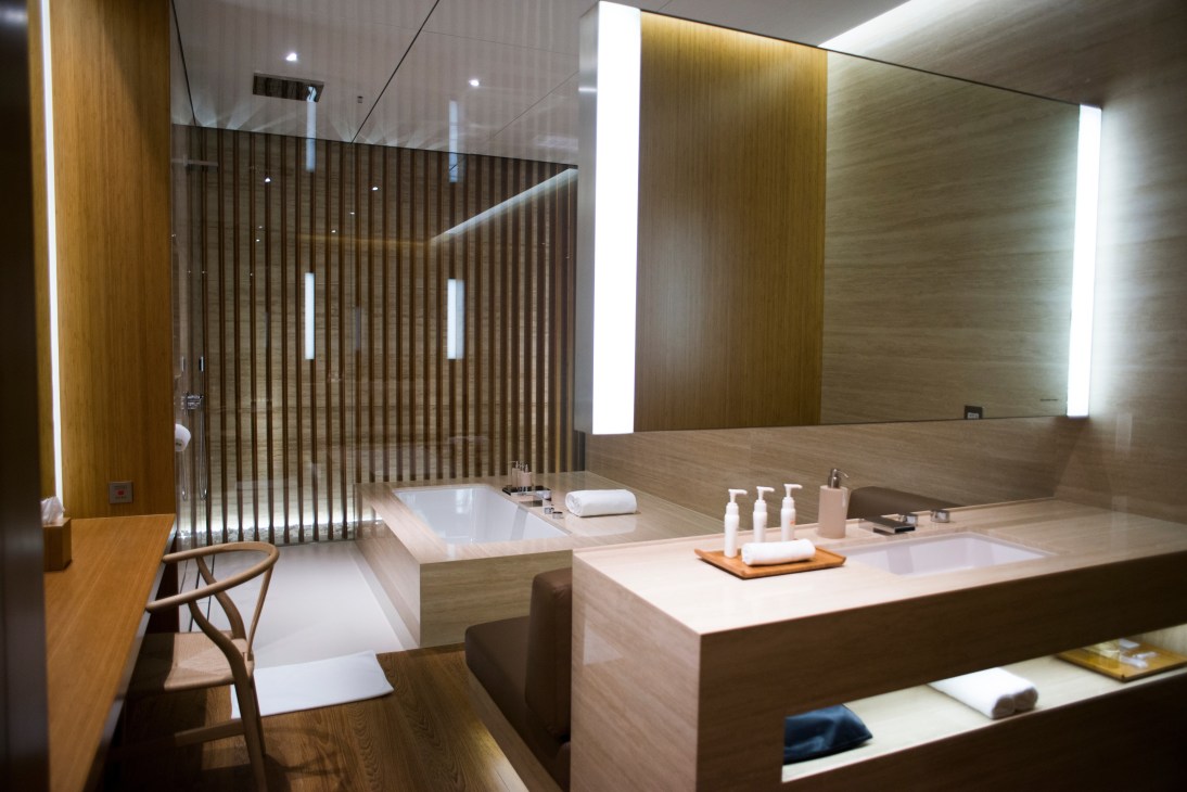 Modern bathroom with wooden finishes, white soaking tub, and illuminated mirror in Cathay Pacific first-class lounge.