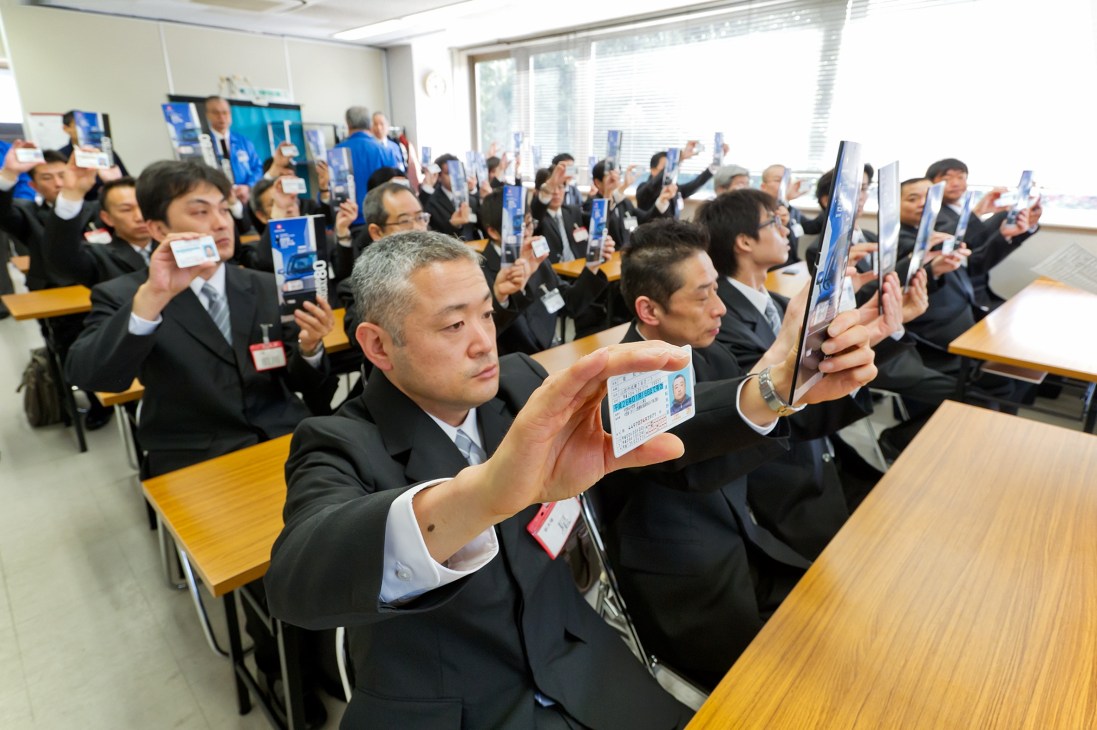 Nihon Kotsu taxi drivers in black suits holding up their licenses during a training session.