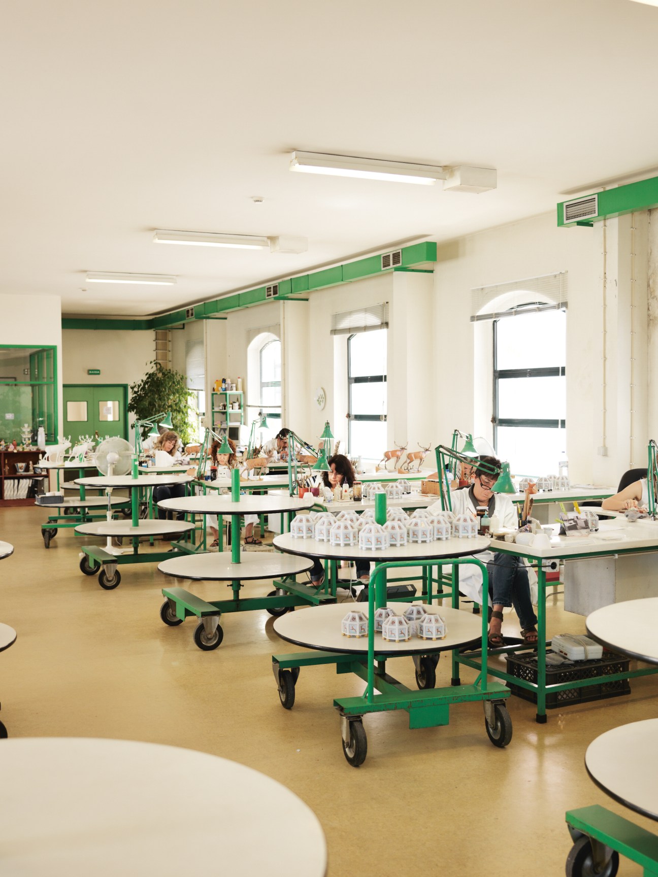 Artists hand-painting white porcelain pieces at round tables in a bright studio with green accents.