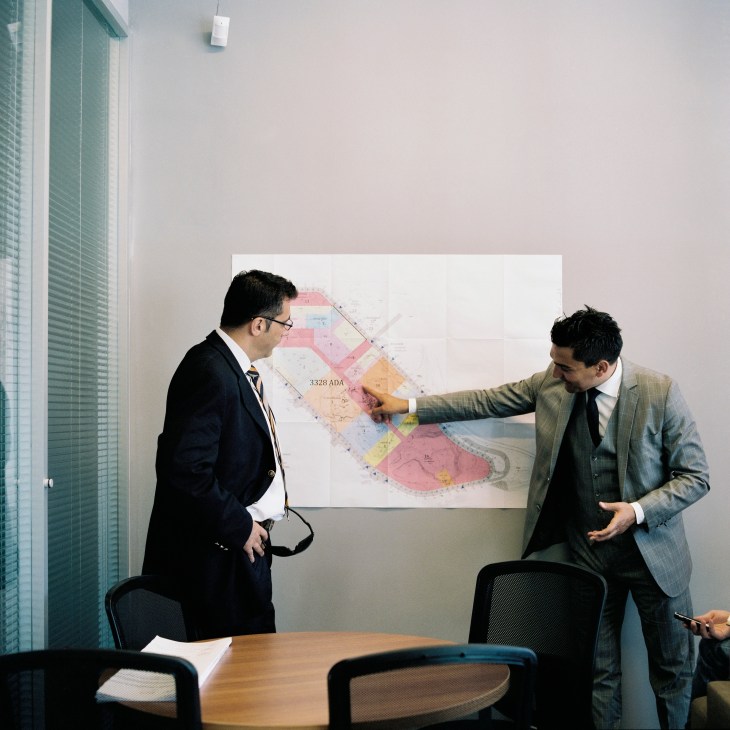 Two men in business suits examine a development plan on a wall in an office setting.