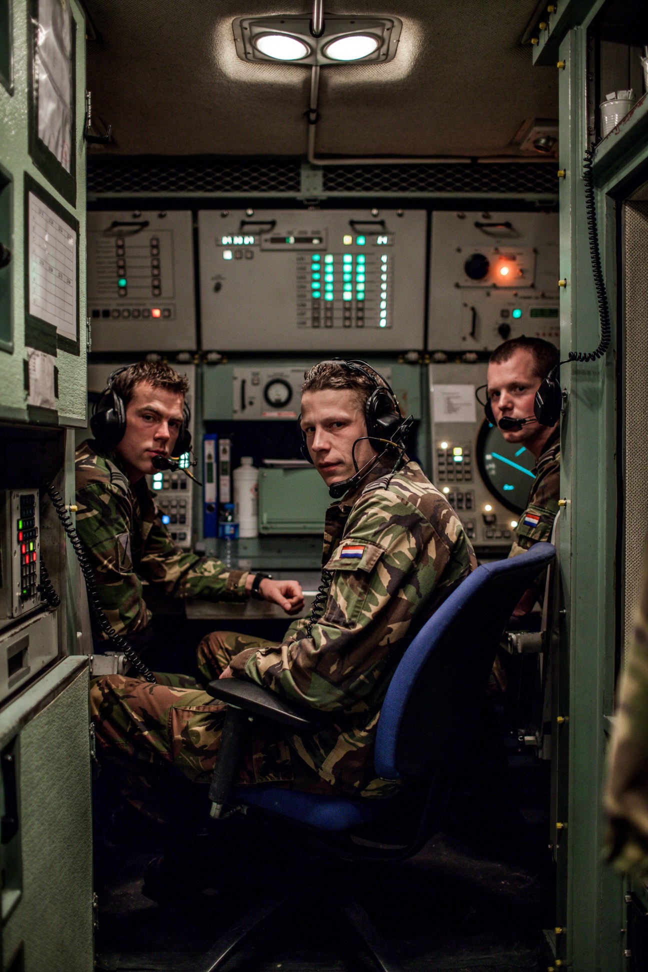 Three Dutch soldiers in camouflage uniforms wearing headsets operate a Patriot missile engagement control station with illuminated control panels.