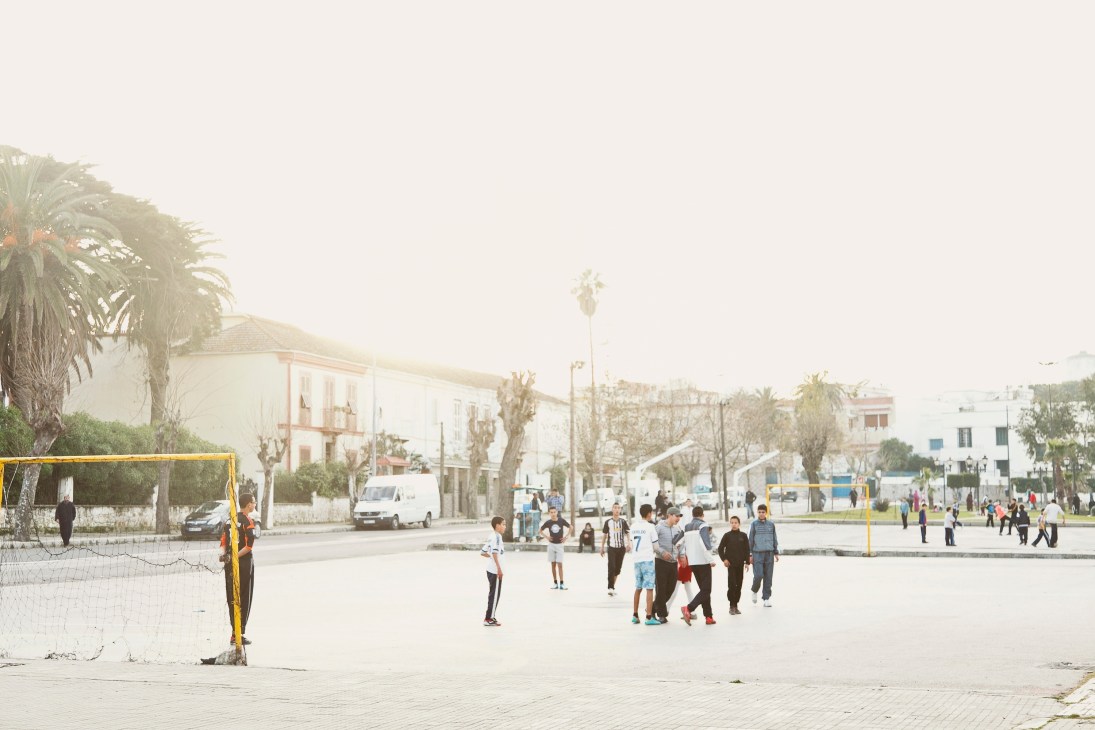 Children playing football in a sunny Marshan square with palm trees and colonial-era buildings in the background.