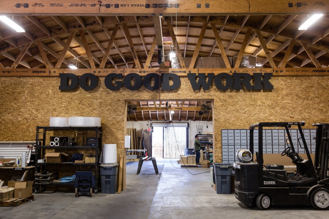 Interior of Woodsmithe workshop showing exposed wooden roof beams, cork walls, and 'DO GOOD WORK' signage above an open production space.