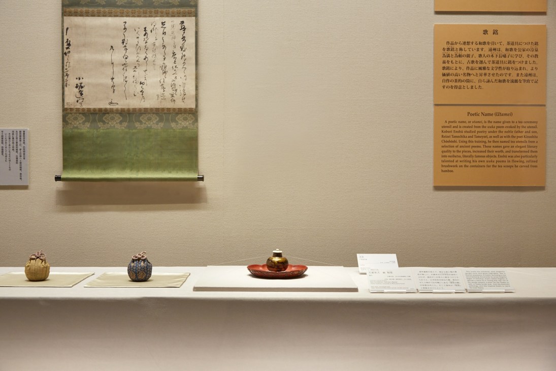 Tea ceremony artifacts displayed in a museum case including ceramic vessels and gold vessels with Japanese calligraphy scroll above.