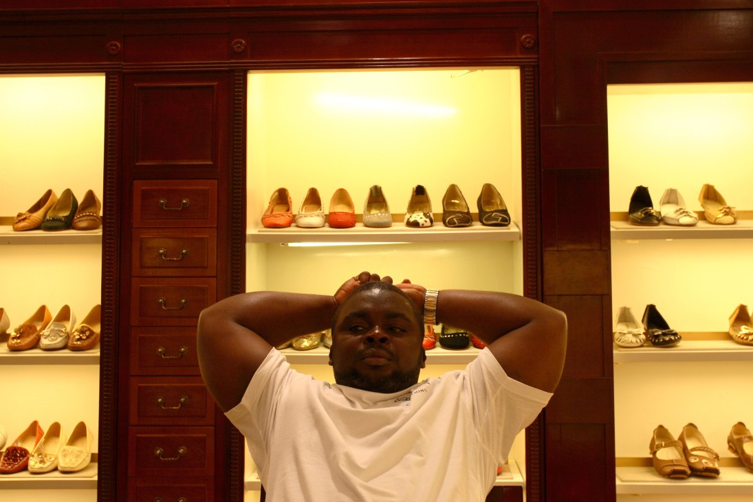 African man relaxing in front of illuminated shoe display shelves in a retail showroom.