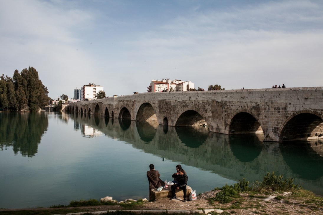 Two people sit by the Seyhan River in front of Adana's historic stone bridge with arches.