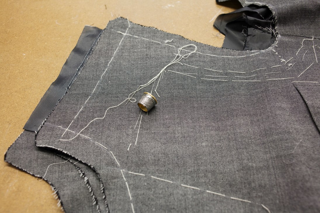 Dark fabric jacket in progress with white tailor's markings and a thimble on a wooden work surface.