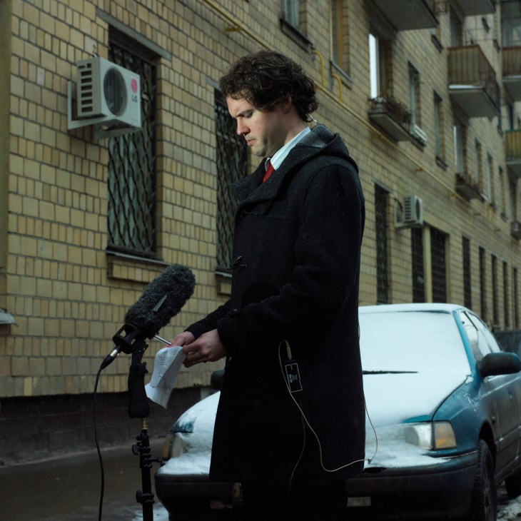 A man in a dark coat holds papers while standing at a microphone on a street with brick buildings.