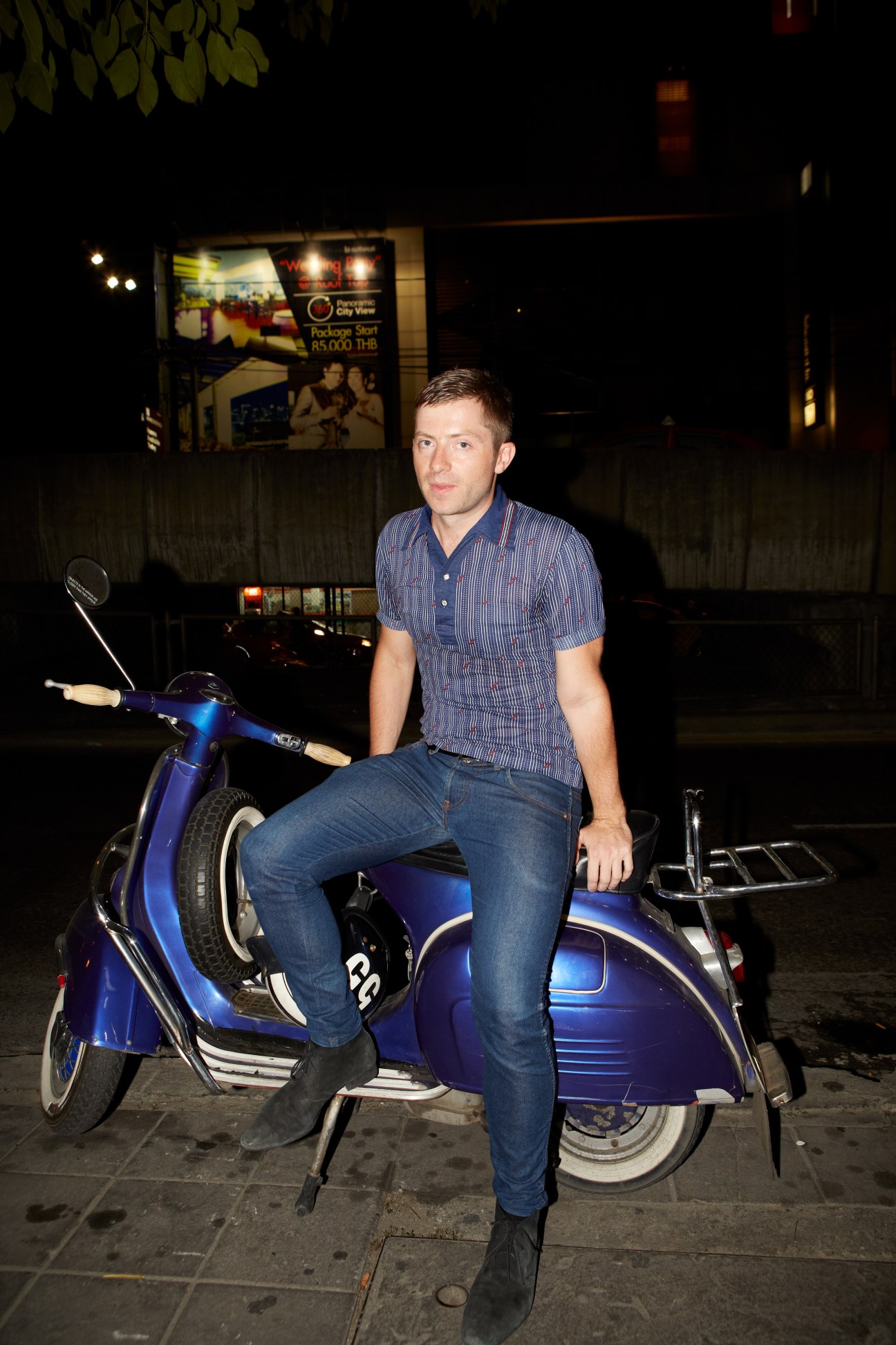 Man sitting on blue Vespa scooter at night on a Bangkok street