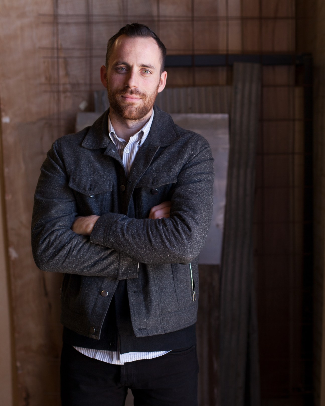 Man in dark jacket and striped shirt standing in industrial workshop with corrugated metal walls.