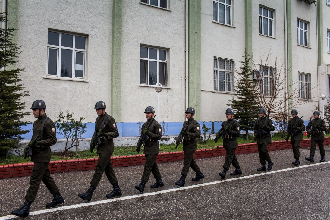 Turkish military police in uniform and helmets stand at ease in a line outside a white and blue building.